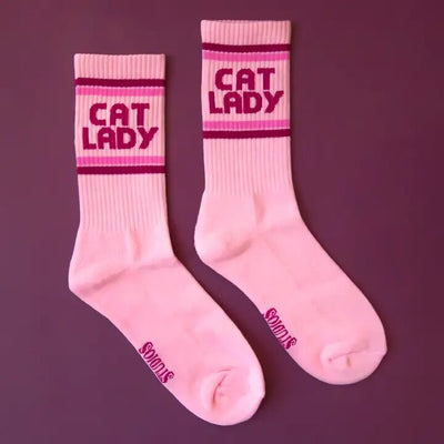 Cat Lady Ribbed Crew Socks are pink with red accents and CAT LADY in bold red text, made by Sunshine Studios. Featuring organic cotton, their name appears near the toes on a solid purple background. Enjoy comfort, style, and eco-friendly packaging.