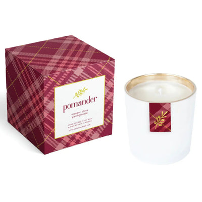 The Annapolis Candle Pomander Boxed Candle features a white candle in a glass holder with a gold rim beside a red plaid box. Its festive scent blends orange, clove, and pomegranate for classic holiday aroma.
