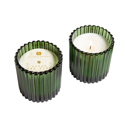Two 7oz. Hillhouse Naturals/Field+Fleur Cedar & Pine candles in green glass show vertical ridges; one is unlit and labeled, the other is lit. Both feature white wax and provide a long burn time.