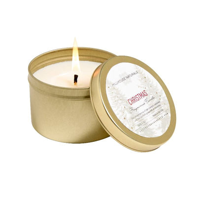 A 5oz Christmas Candle in a gold tin from Hillhouse Naturals/Field+Fleur, shown lit. The lid leans against the tin, displaying a festive white and gold CHRISTMAS label with tree motifs and details about its long burn and fragrance.