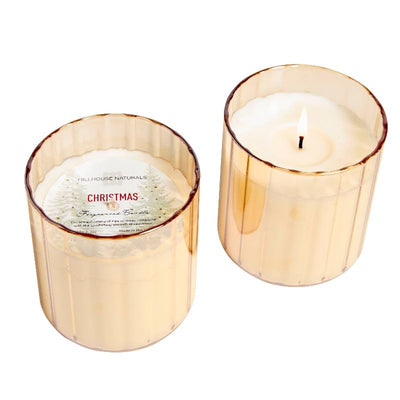 The Hillhouse Naturals/Field+Fleur 7oz Christmas Candle in a small gold glass offers festive aroma and long burn time. One candle is lit; another, lid off, shows a CHRISTMAS label and pine tree illustration—perfect for holiday decor.