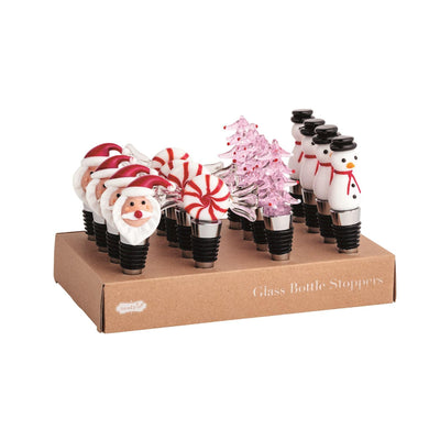 The Mud Pie Christmas Glass Bottle Stoppers display box holds metal stoppers topped with glass Santa faces, peppermint candies, pink Christmas trees, and snowmen, all arranged in four neat rows.