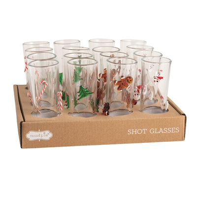 Mud Pie’s Christmas Shotglasses set includes twelve clear glasses in a cardboard tray, each adorned with festive designs—candy canes, trees, gingerbread men, and snowmen—and charming dimensional details.