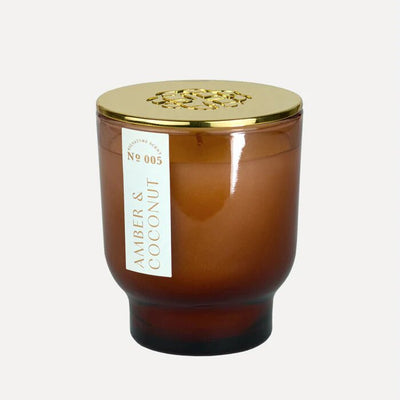 The Amber & Coconut 7 oz Maeve Glass Candle by Elum features a brown glass jar with a gold lid, embossed floral design, labeled Amber & Coconut No 005. It highlights its soy wax and Aura Pure technology for an elegant touch.