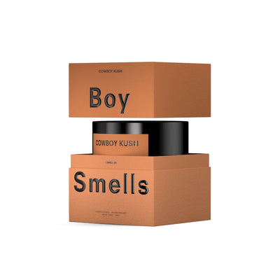 A minimalistic brown box labeled Boy Smells holds the Cowboy Kush Hero Candle. The top and bottom are separated, revealing the black candle inside with bold black text on the packaging.
