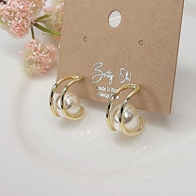 The Cradled Pearl Hoop Earrings by BETTYOH INC feature large white pearls on gold-tone hoops with titanium posts, displayed on a card reading Betty Oh! Made in Korea. Lead and nickel free for sensitive ears. Pink flowers decorate the background.
