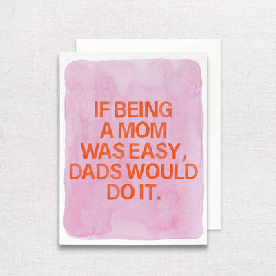 The If Being a Mom Was Easy, Dads Would Do It Greeting Card by Gert & Co, a woman-owned brand, showcases bold orange text on a pink watercolor background. Made from recycled paper, it elegantly complements any textured, light gray surface—an ideal Mothers Day card.