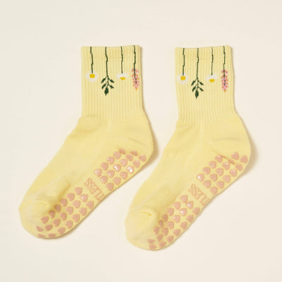 The Effortless Club Flower Stem Grip Sock is a yellow Pilates sock with embroidered hanging flowers at the cuffs, a cushioned footbed, and pink non-slip grips on the soles.