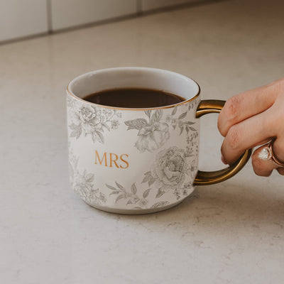 A hand with a ring holds the elegant Mrs Coffee Mug by Sweet Water Decor, featuring gold accents and floral designs. Filled with coffee, this fine bone china mug is the perfect bridal gift for any special occasion.