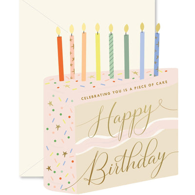 The Ginger P. Designs Piece of Cake die-cut folded Greeting Card features a cake shape with seven lit candles, colorful sprinkles, and gold foil accents. The card reads Happy Birthday and Celebrating you is a piece of cake. Envelope included.
