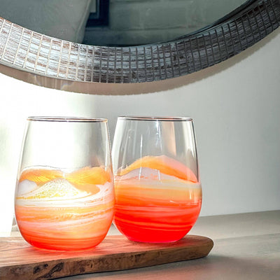 Two Artful Nests Resin Wave Sunset Stemless Wine Glasses, filled with a vibrant orange and white drink, sit on a wooden board before a round textured mirror, as sunlight casts reflections across the scene.