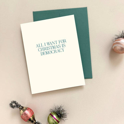 The "All I Want For Christmas Is Democracy Letterpress Card" by Missive features bold text on FSC cardstock, displayed with festive ornaments and a green envelope atop a light surface.