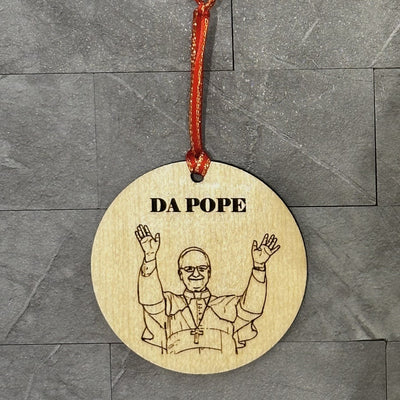 The DA POPE Ornament by Jim Harris is a 3-inch birchwood hanging piece, laser etched with a figure in religious attire raising both hands and "DA POPE" above, hung by a red ribbon against gray tiles.