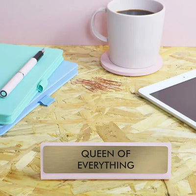 The Flamingo Candles Queen of Everything Deskplate, in pink and gold engraving, is the perfect motivational gift. Nearby are a mug on a coaster, a pen atop blue-green notebooks, scattered paperclips, and a tablet on the wooden desk.