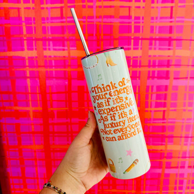 A hand holds the Sonny Rising "Your Energy Is Expensive" 20 oz Skinny Tumbler with straw in front of a pink and orange checkered wall. The tumbler features vibrant lettering reminding you your energy is a luxury not everyone can afford.
