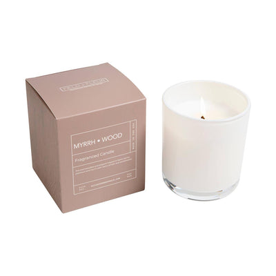 A Hillhouse Naturals Field+Fleur Myrrh Wood Candle in white glass is lit beside its light brown box. The 9oz candle offers soothing myrrh and sandalwood notes, with details in white text on the MYRRH + WOOD labeled packaging.