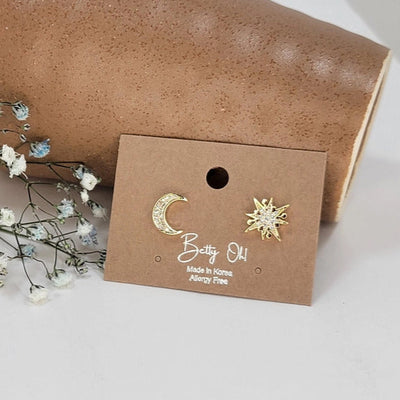 A brown card displays BETTYOH INCs Star and Moon Mismatched Stud Earrings—one crescent moon, one starburst—with Allergy Free and Made in Korea printed on it. White flowers and a brown cylinder appear in the background.