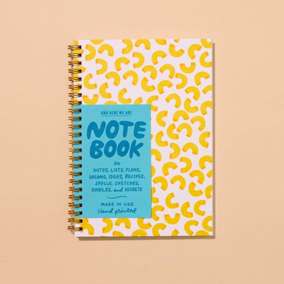 The Macaroni Letterpress Spiral Notebook by And Here We Are is an A5, spiral-bound notebook with a white cover featuring yellow macaroni shapes and a blue “NOTEBOOK” label—ideal for lists, plans, recipes, or pasta-inspired doodles.