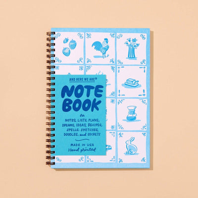 The And Here We Are Delft Tile A5 Letterpress Spiral Notebook is spiral-bound with a blue and white cover inspired by delft tiles, featuring hand-drawn illustrations of fruits, a rooster, vase, book, coffee pot, and rabbit.