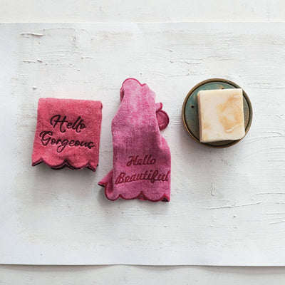 The Creative Co-op Hello Gorgeous & Hello Beautiful Cotton Terry Washcloths - Set of 2 are pink with scalloped edges and embroidered phrases, shown next to a soap bar on a small dish atop a white surface.