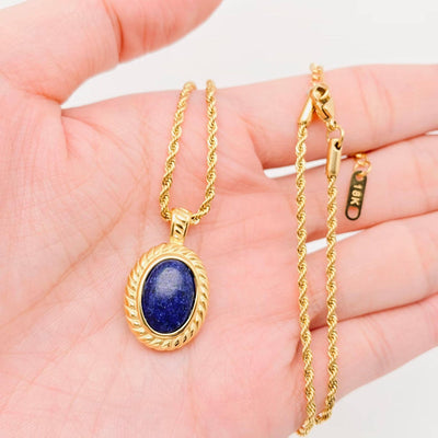 A hand holds the Mio Queena Lapis Lazuli 18K Gold-plated Steel Pendant Necklace—an oval deep blue stone set in a twisted 18K gold-plated stainless steel frame, with a tag showing its 18K gold content.