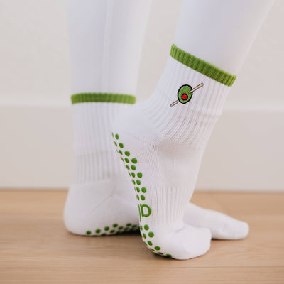 A person stands tiptoe on light wood flooring, wearing Almla Active's Olive You Pilates Socks—women’s cotton socks with green cuffs, embroidered olive graphic, green non-slip dots, and part of a green "p" on the soles.
