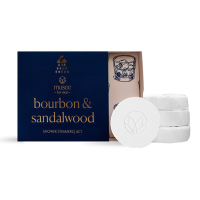 A dark blue Musee box labeled Bourbon & Sandalwood Shower Steamers sits next to three white steamers, its lid partially open to reveal an illustration of a bourbon glass and a touch of sandalwood essential oil inside.