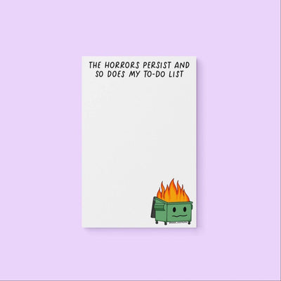 The Horrors Persist Notepad by MangoIllustrated features "The horrors persist and so does my to-do list" atop a lavender sheet with a cheerful dumpster fire cartoon—perfect for adding humor to productivity and burnout.