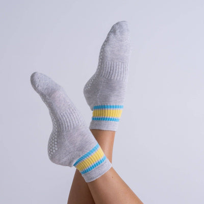 Two feet in Reformer Loops’ Cali Quarter Pilates Grip Socks—grey with yellow and blue stripes—are crossed on a plain light gray background. White silicone grips on the soles provide added traction.