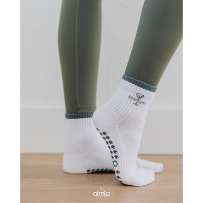 A person in olive green leggings and Almla Active Extra Dirty Pilates Socks with grip soles stands on wooden flooring, heels raised. The white socks display “extra dirty” and a martini glass design. Brand: Almla Active.