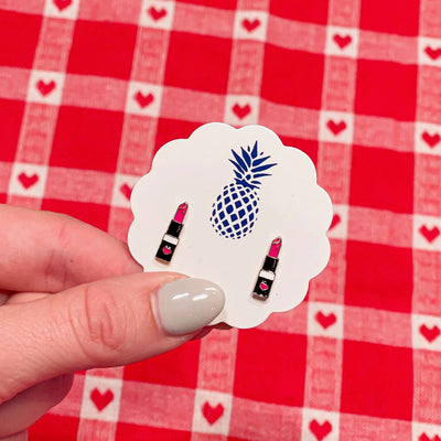 A hand with light gray nail polish holds a scalloped white card featuring a blue pineapple, showcasing Prep Obsessed Wholesale Lipstick Enamel Stud Earrings, against a red and white checkered heart-accented tablecloth.