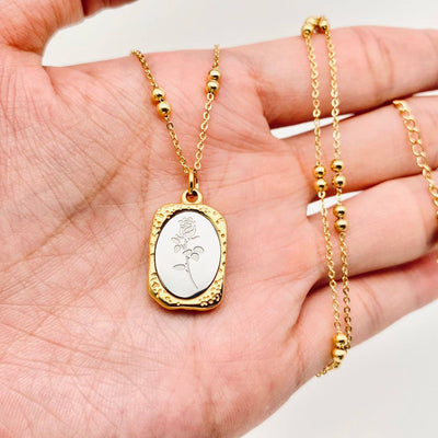 A hand holds the Mio Queena Magic Mirror Rose Stainless Steel Pendant Necklace, featuring an oval 18K gold plated pendant with an engraved rose, textured gold border, and a delicate chain adorned with small gold beads.