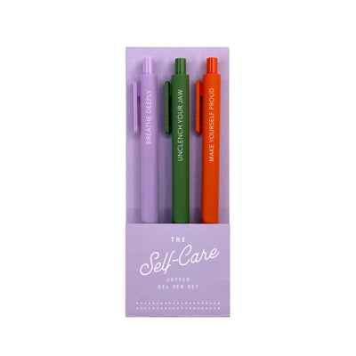 The Ruff House Print Shop Self-Care Jotter Gel Pen Set of 3 includes purple (“BREATHE DEEPLY”), green (“UNCLENCH YOUR JAW”), and orange (“MAKE YOURSELF PROUD”) pens in a colorful purple box.
