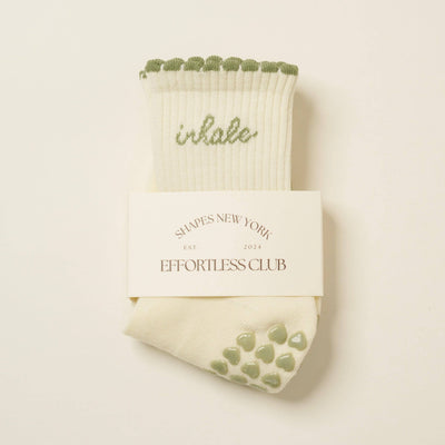 The Inhale Exhale Grip Sock by Effortless Club is a cream-colored organic cotton sock with green trim, inhale stitched near the top, and green heart-shaped grips on the sole—perfect for Pilates.