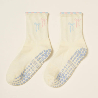 The Effortless Club Ribbon Duo Petal Grip Sock is a cream organic cotton pair featuring blue and pink bows near the top, scalloped cuffs in matching shades, cushioned footbeds, and blue non-slip dots on the soles.