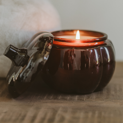 The Sweet Water Decor Hello Fall Soy Candle - 20oz Pumpkin Jar, glowing with a warm flame and autumn scent, sits on a wooden surface, creating a cozy and inviting fall decor ambiance.