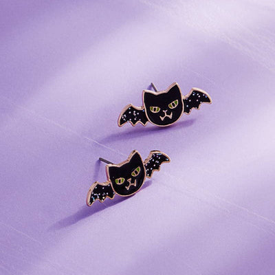 The Halloween Batcat Enamel Titanium Post Earrings by Designature New York are hypoallergenic studs shaped like black cats with bat wings, yellow eyes, and silver stars, shown on a light purple background.