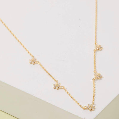 The Fame Accessories Dainty CZ Flower Station Necklace features small flower-shaped cubic zirconia pendants, delicately spaced along the chain and elegantly displayed on a white geometric surface.