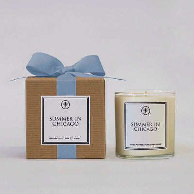 The Summer in Chicago 11 oz candle by Ella B Candles is presented in a glass jar with jasmine notes, packaged in a brown box tied with a light blue ribbon. Both the jar and box have white labels with black text and an illustration.