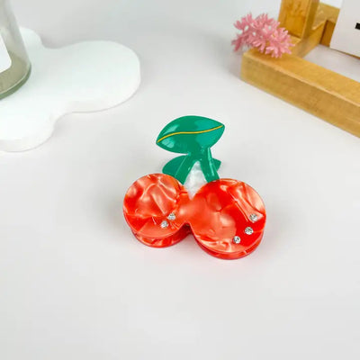 The mildstyles Cherry Hair Claw Clip with Rhinestones, featuring two red cherries with green leaves, rests on a white surface beside a wooden tray and a pink artificial flower.