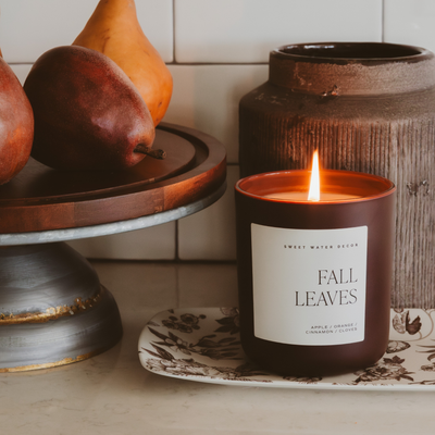 The Fall Leaves 15 oz Candle in Matte Jar by Sweet Water Decor sits on a decorative plate beside a cake stand with pears and a rustic brown jar, creating a cozy autumn vibe on the kitchen counter.