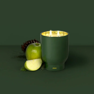 An Elum Designs Home Pine Birch 21 oz Maeve Glass Candle in a glossy holder glows beside a green apple, slices, pine needles, and a pine cone on dark green, its golden interior reflecting the flame and filling the air with its crisp aroma.