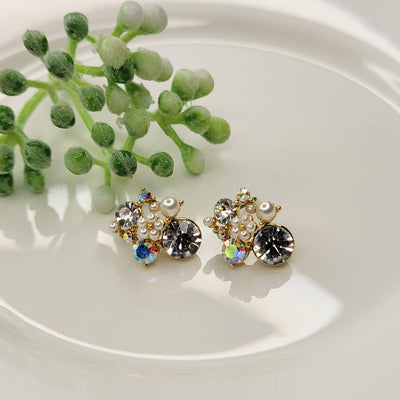 The Seraphina Post Earrings by BETTYOH INC feature decorative rhinestones, pearls, and iridescent gemstones in a cluster design. Lead and nickel free, they are gentle on sensitive ears. Shown on a white surface with a green plant backdrop.