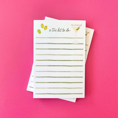 The Gert & Co A Tini Bit To Do Martini Notepad (4x6) features green lines, olive and martini glass art, and sits on a pink background with another to do list pad partially visible underneath.