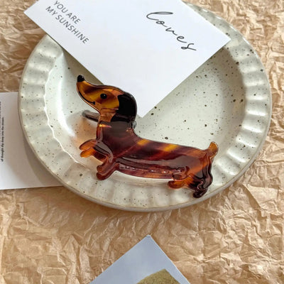 The SoapElenGlen Dachshund Hair Claw Clip with a tortoiseshell pattern sits on a textured white plate, alongside decorative cards, crumpled beige paper, and lightweight earrings.