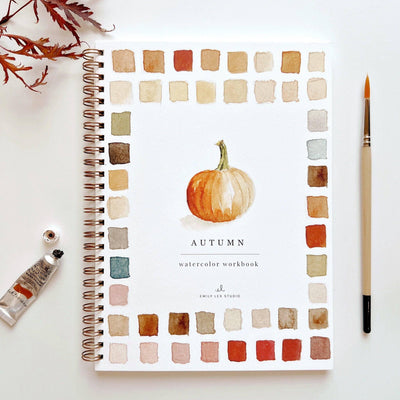 The emily lex studio Autumn Watercolor Workbook is spiral-bound and features a painted pumpkin center with fall watercolor art. A paintbrush and watercolor tube rest beside it on a white surface.