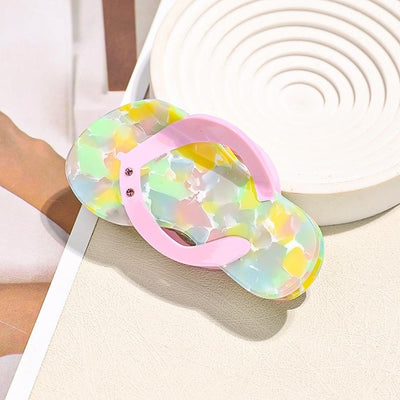 A vibrant, translucent Acetate Flip Flop Claw Hair Clip by Judson & Company features pink straps and a sole showcasing yellow, green, and peach tones. The backgrounds spiral design adds texture to the scene.