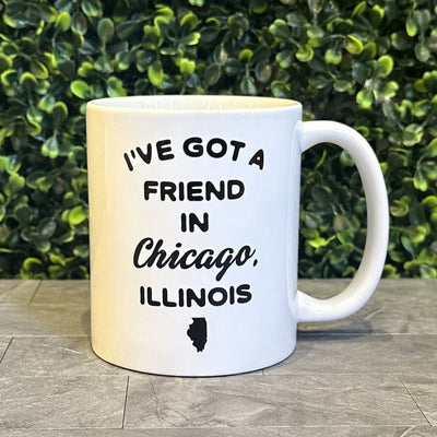 The Mugsby "I've Got A Friend In Chicago" 11 oz mug is white ceramic with black text and a state silhouette, dishwasher and microwave safe, shown on a gray surface with green foliage in the background.