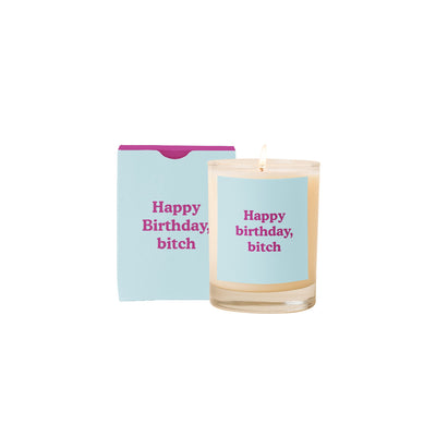 The Birthday Bitch Candle by Talking Out of Turn features a white soy-based wax candle in a glass holder with Happy birthday, bitch in pink. A matching box with the same text and purple top sits behind, while the lit candle casts a warm glow on the white backdrop.
