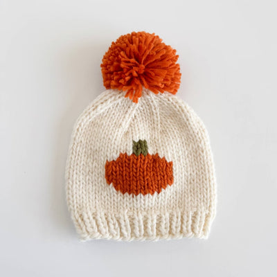 The Figgy Friends Pumpkin Knit Baby Hat is a white hand-knit hat for babies, with a large orange pom-pom and pumpkin design—perfect for fall.